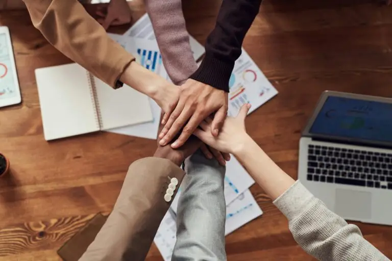 Team of Excel experts joining hands over a desk with business graphs, spreadsheets, and an open laptop